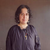 Headshot of Alexander Te Pohe. He is wearing a loose black shirt, a sword necklace, and white petal earrings. His hair is almost shoulder length, black, and wavy. He is smiling a little.